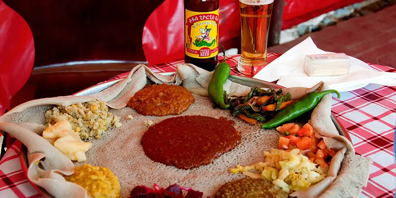 Tradycyjna kuchnia etiopska na płaskim chlebie injera, z różnymi gulaszami, warzywami i papryczkami. Obok piwo.