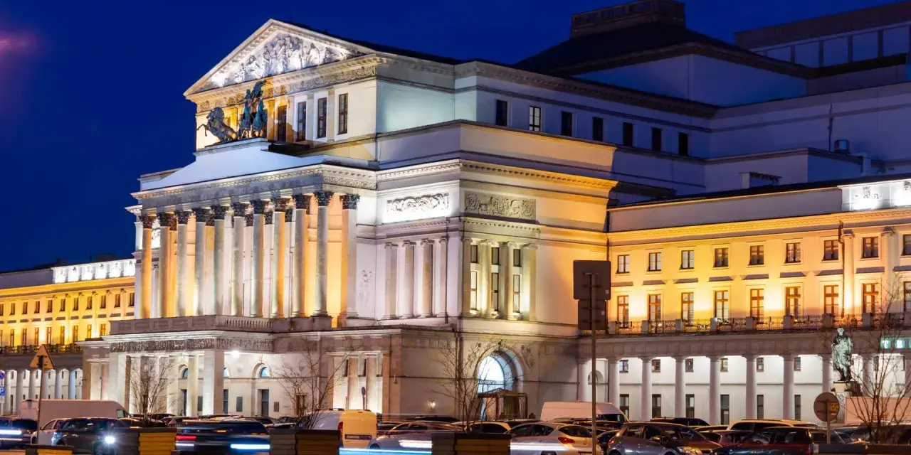 Teatr Wielki w Warszawie, najstarszy teatr w Polsce, nocą. Monumentalna fasada z kolumnami i rzeźbami, oświetlona ciepłym światłem, z samochodami w ruchu ulicznym przed budynkiem.