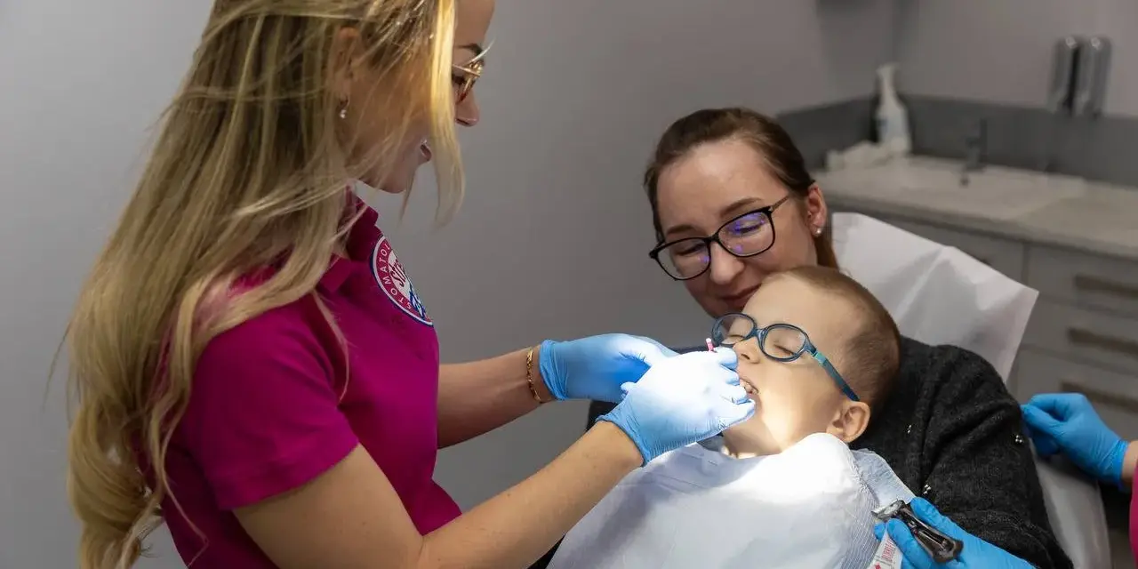 Dentystka bada zęby chłopca, podczas gdy jego mama go wspiera. Zwracają uwagę na zdrowie dziąseł, by zapobiec recesji dziąseł.
