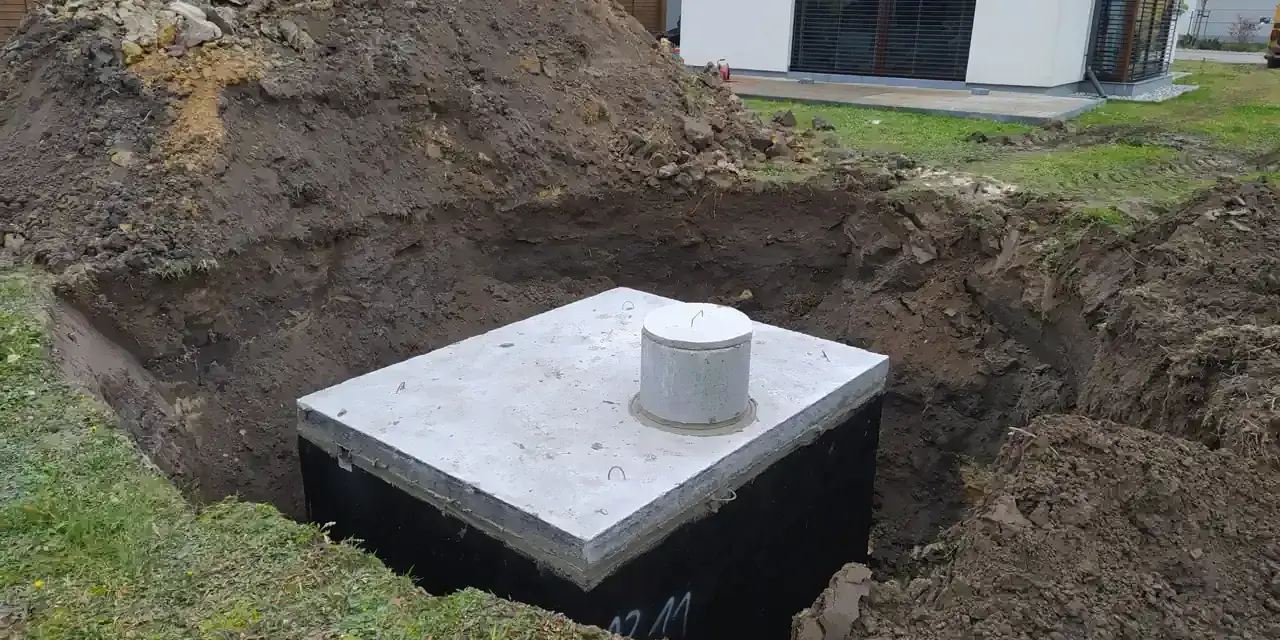 Gotowy odpływ z rynny do ziemi, betonowy zbiornik w wykopie, obok hałda ziemi i nowoczesny dom.