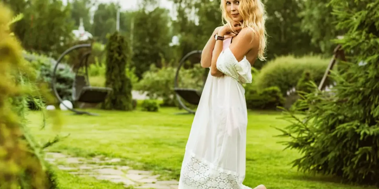 Uśmiechnięta kobieta panna w zwiewnej białej sukni, z rozpuszczonymi blond włosami, stoi na trawniku w ogrodzie.