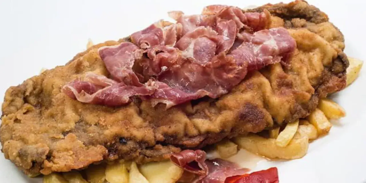 Cachopo asturiano rebozado con jamón serrano encima, acompañado de patatas fritas y pimientos rojos.
