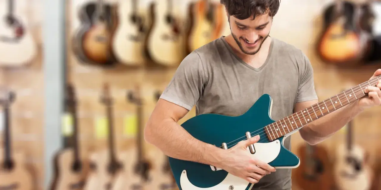 Uśmiechnięty mężczyzna testuje gitarę elektryczną w sklepie muzycznym. W tle widać wiele innych instrumentów.