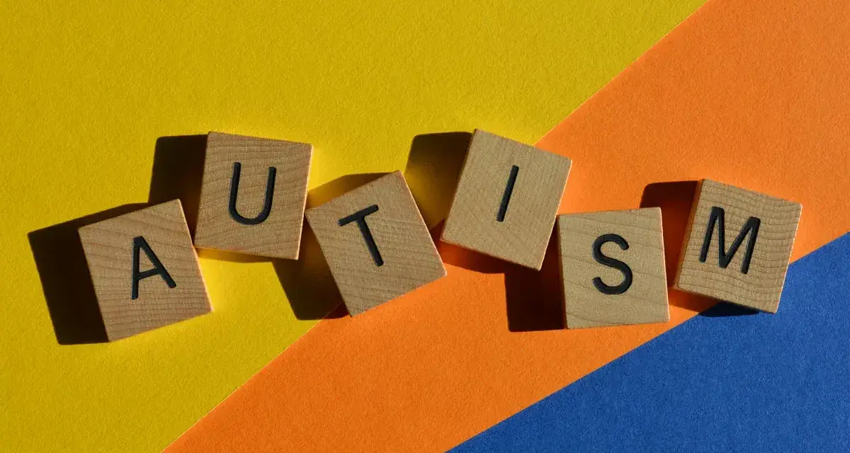 Lettere di legno formano la parola AUTISM su uno sfondo colorato. La domanda "autistici si nasce o si diventa" risuona.