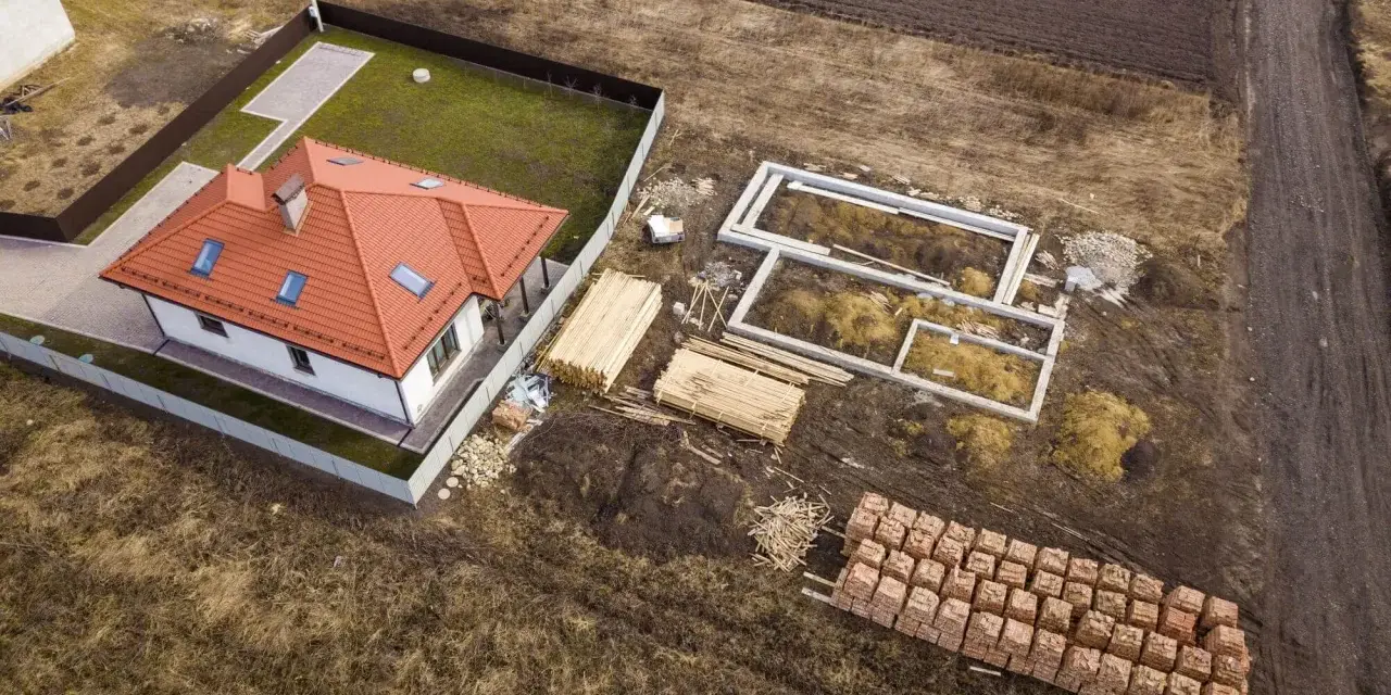 Zabudowa zagrodowa w budowie: gotowy dom z czerwonym dachem, fundamenty, drewno i palety cegieł na placu budowy.
