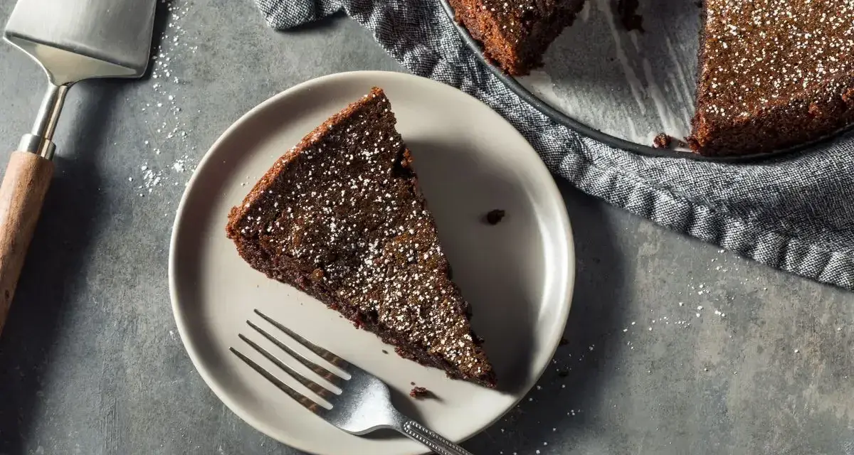 Una fetta di torta all'acqua al cacao, spolverata di zucchero a velo, su un piatto con una forchetta. Accanto, la torta intera e una paletta.