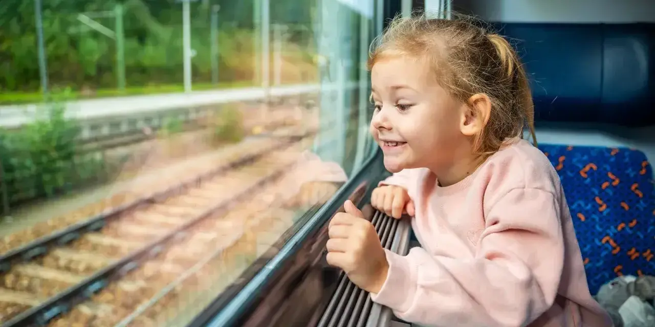 Uśmiechnięta dziewczynka patrzy przez okno pociągu. Zastanawia się, od ilu lat można jeździć samemu pociągiem, marząc o podróżach.