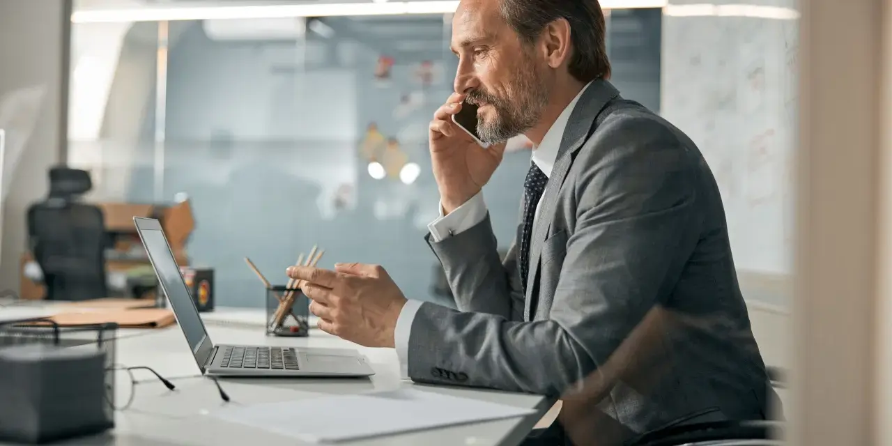 Biznesmen w garniturze rozmawia przez telefon, pracując przy laptopie. To typowy dzień w życiu CEO, co to znaczy być liderem.
