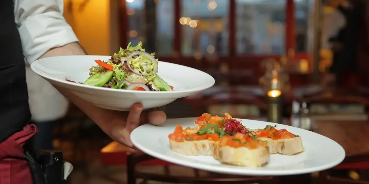 Un camarero experto demuestra cómo servir una mesa, presentando un plato de ensalada fresca y bruschettas.