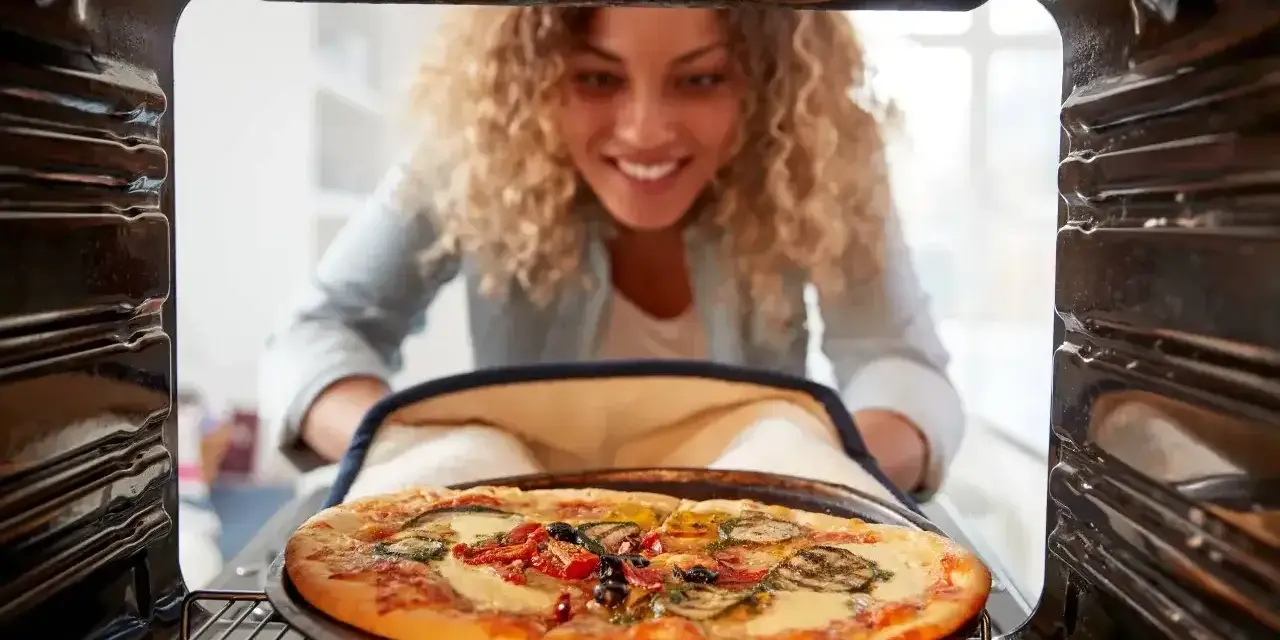 Uśmiechnięta kobieta wyjmuje z piekarnika pyszną, gorącą pizzę. Właśnie skończyła piec pizzę i jest gotowa do jedzenia.