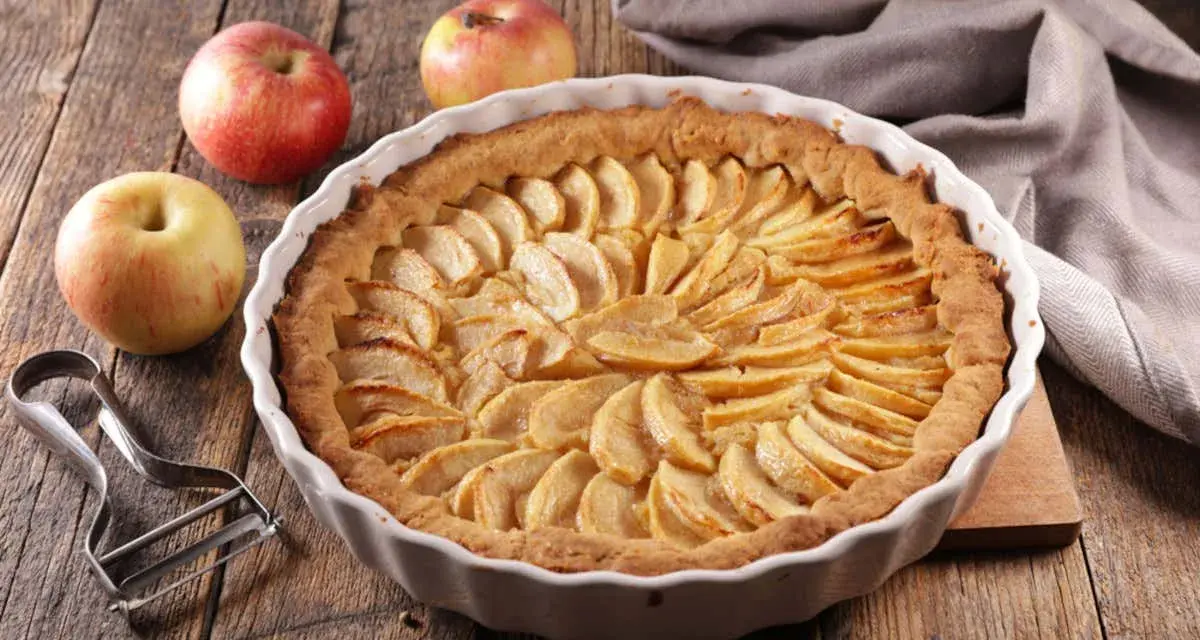 Torta di mele umida e soffice, con mele fresche e un pelapatate su un tavolo di legno rustico.