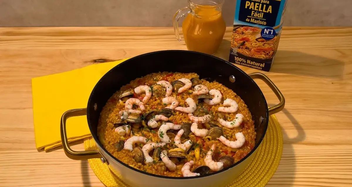 Delicioso arroz a la marinera en paellera, con gambas y mejillones. Al lado, un cartón de caldo para paella de marisco y una jarra con líquido naranja.
