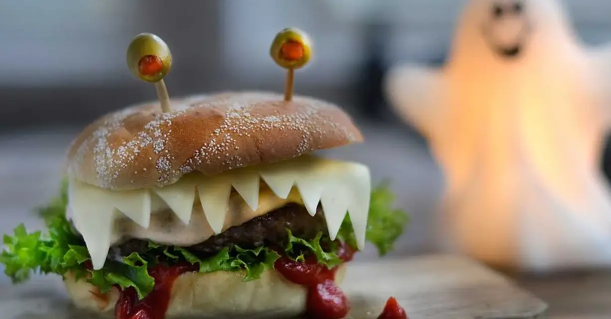 En läskig halloween hamburgare med osttänder och olivögon. En spöklampa lyser upp i bakgrunden.