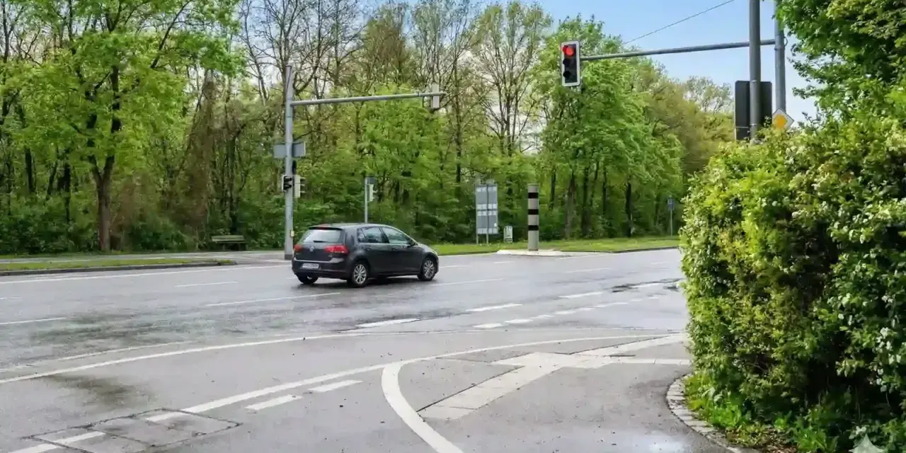Ein dunkles Auto fährt auf nasser Straße an einer roten Ampel vorbei, neben der ein ampel blitzer steht.