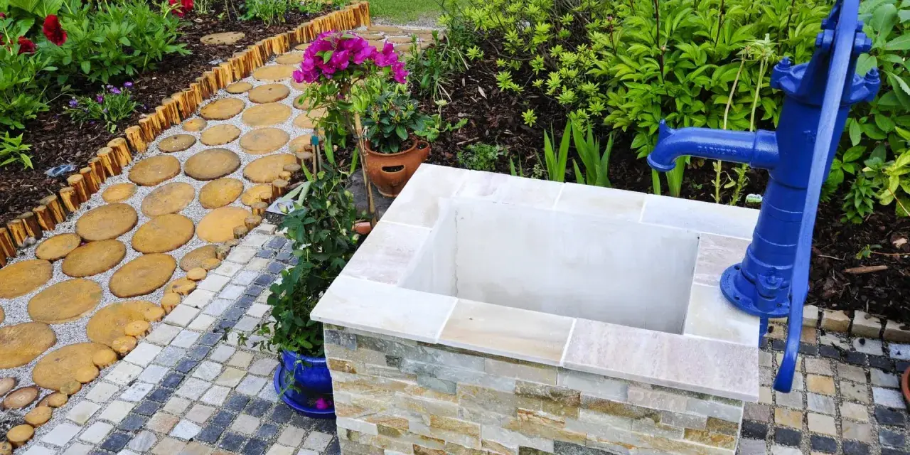 Ein Gartenweg führt zu einem frisch gebauten Brunnen mit blauer Handpumpe.