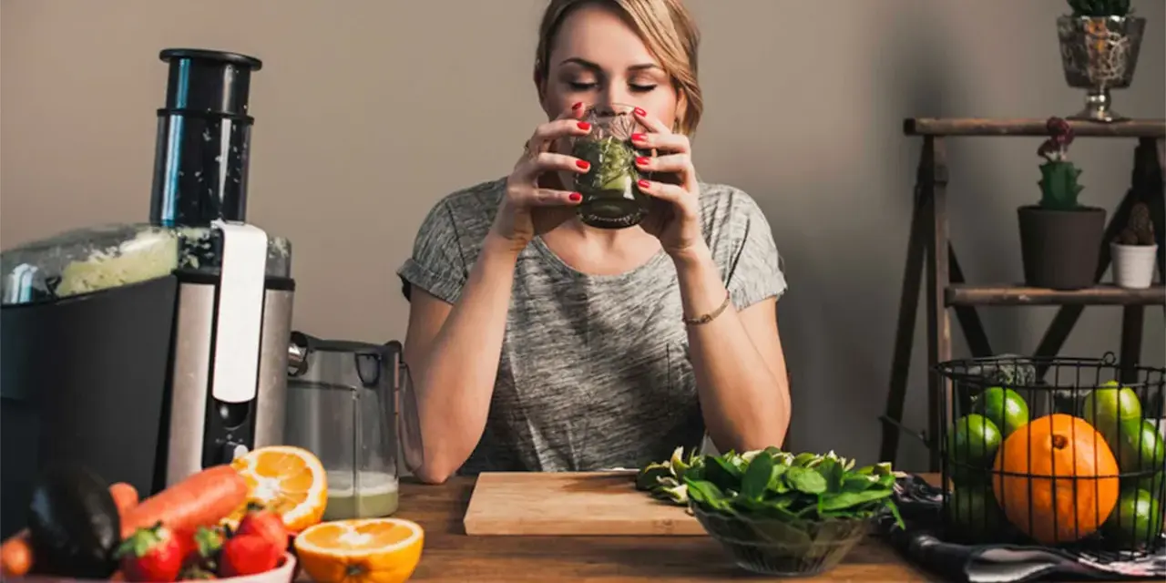 Kvinna dricker grön juice, omgiven av färsk frukt och grönsaker. Hon verkar njuta av sin hälsosamma dryck, kanske som en del av hur deffar man.