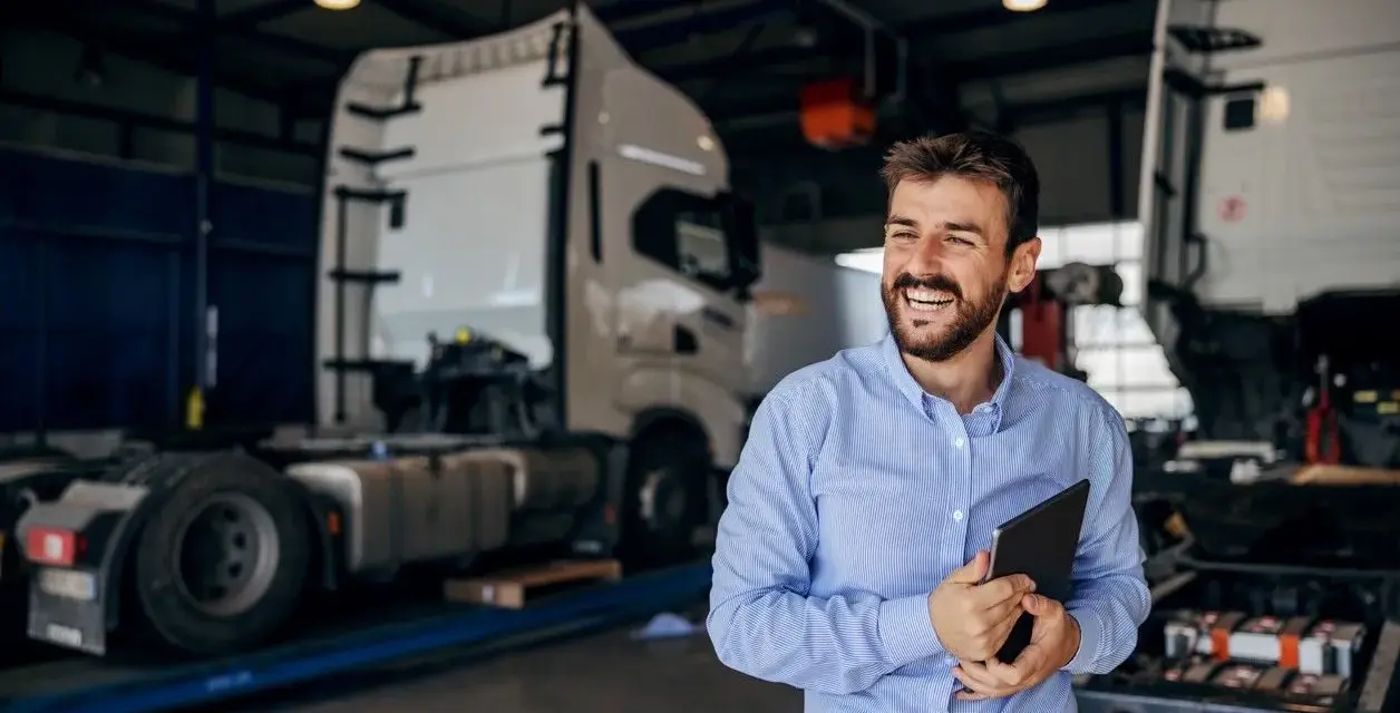 Uśmiechnięty mężczyzna z tabletem w ręku, w tle warsztat z ciężarówkami. Oferuje **usługę transportową ryczałt**.