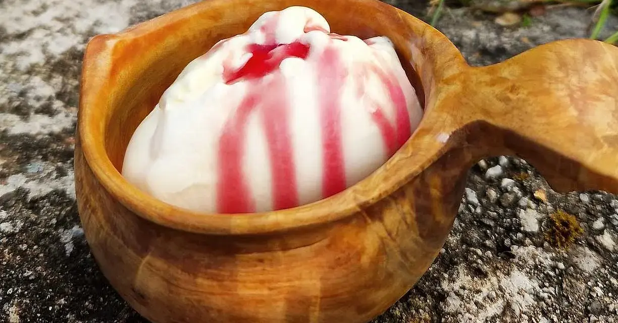 En skopa hemmagjord glass med röd sås i en träkopp, placerad utomhus på sten.