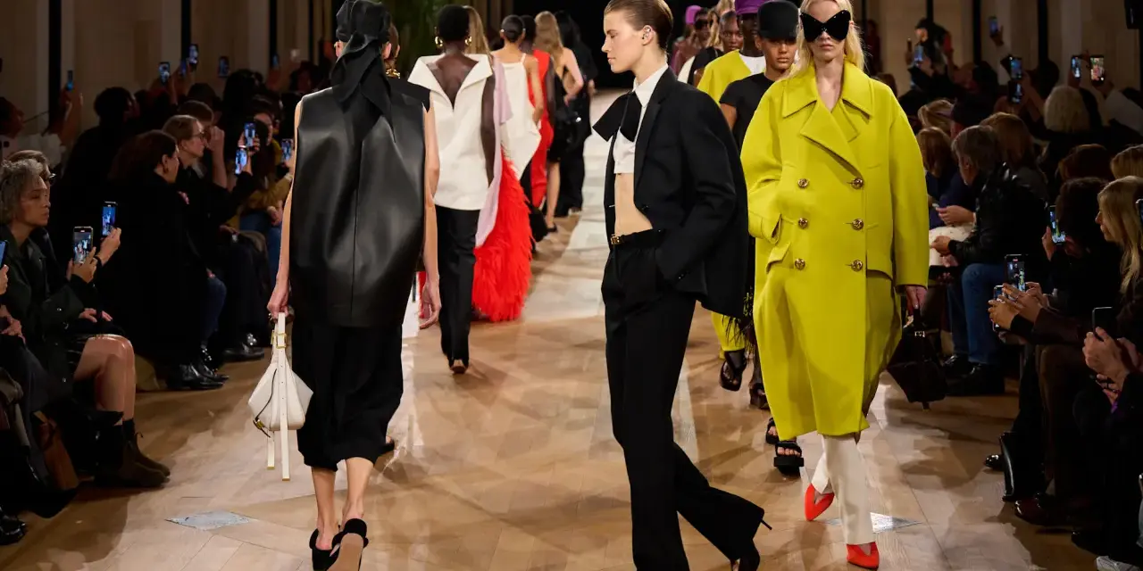 Sfilata Balenciaga: modelli con abiti audaci, tra cui un cappotto giallo acceso e un completo nero, sotto gli occhi del pubblico.