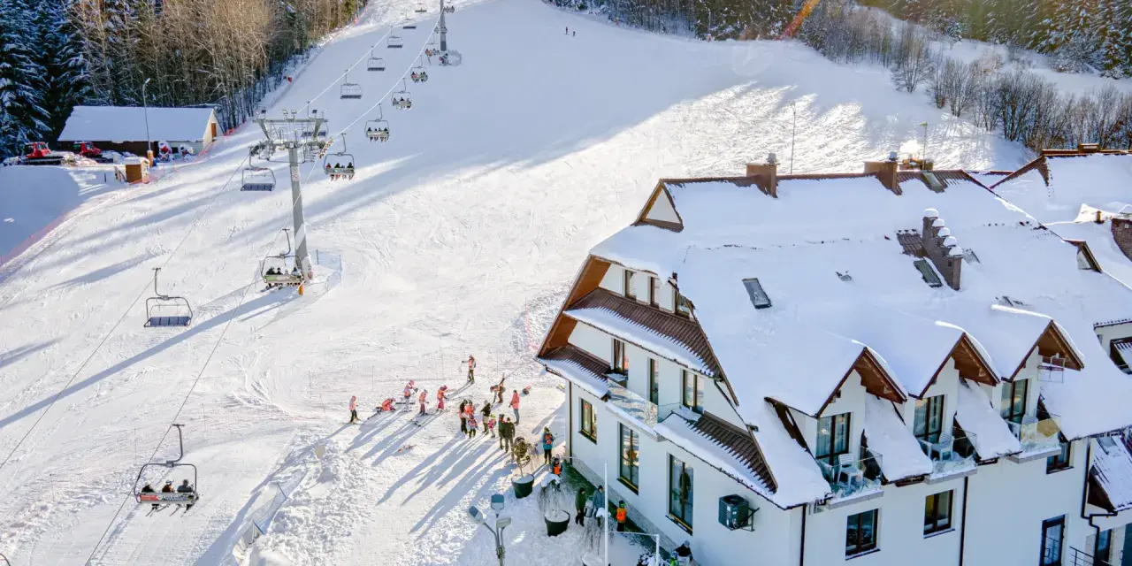 Hotel ze stokiem narciarskim dla dzieci. Kolejka krzesełkowa wwozi narciarzy na ośnieżony stok, a obok hotelu bawią się dzieci.