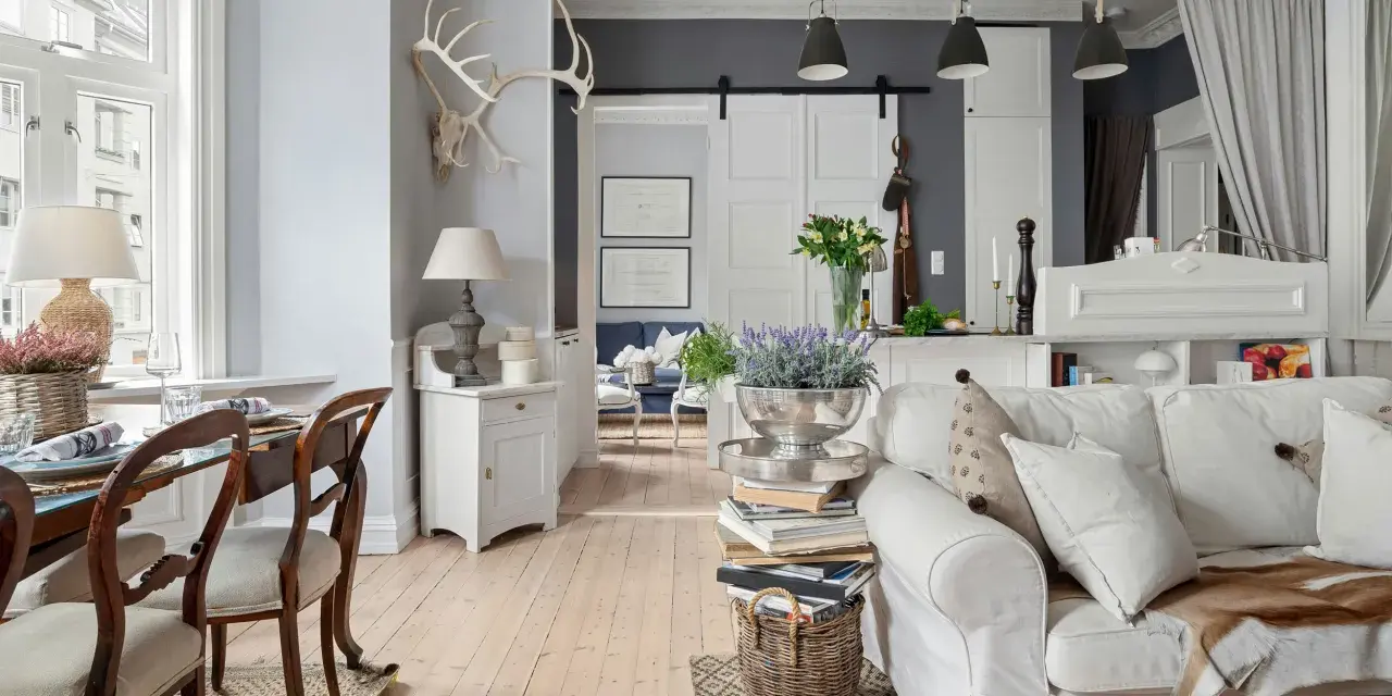 Un salotto con un'atmosfera da arredamento francese chic. Tavolo da pranzo in legno, divano bianco accogliente, lampade e decorazioni rustiche.