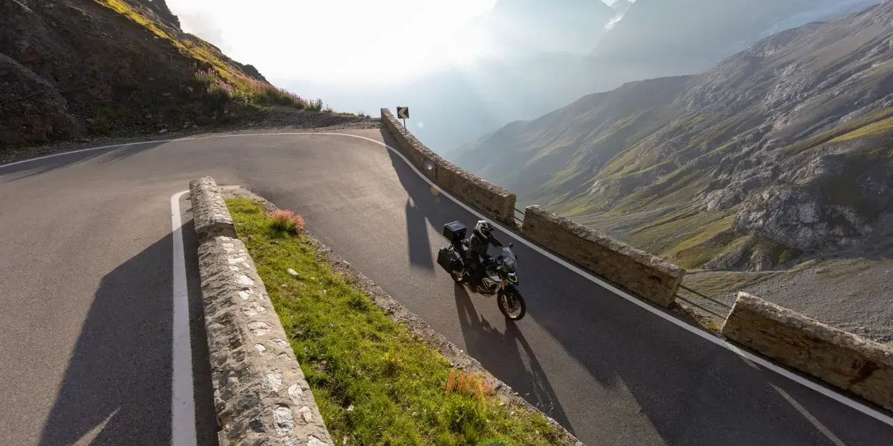Motocyklista przemierza serpentyny górskiej drogi, odkrywając najpiękniejsze trasy motocyklowe w Europie. Słońce przebija się przez mgłę, oświetlając majestatyczne szczyty.