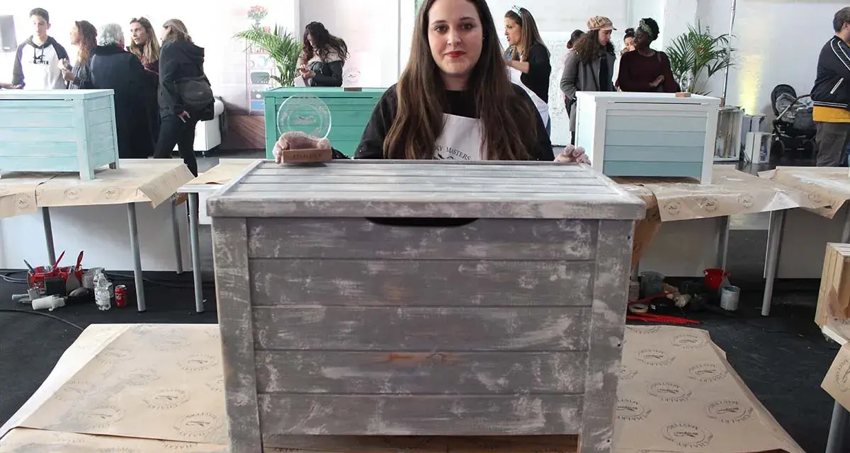 Una mujer muestra un baúl pintado a la tiza con un acabado desgastado. Otros baúles decorados se ven en el fondo.