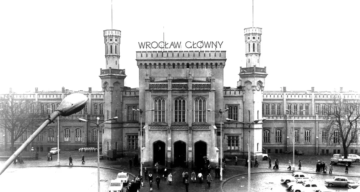 Wrocław Główny, imponujący dworzec z wieżami i zegarem, otoczony samochodami i ludźmi.