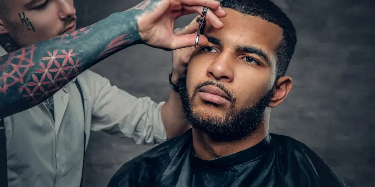 Barbero con tatuajes en el brazo recorta las cejas de un hombre con barba, creando un look buzz cut.