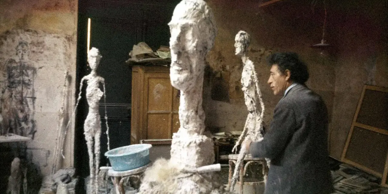 Alberto Giacometti w pracowni, otoczony swoimi charakterystycznymi, smukłymi rzeźbami i niedokończonymi formami.