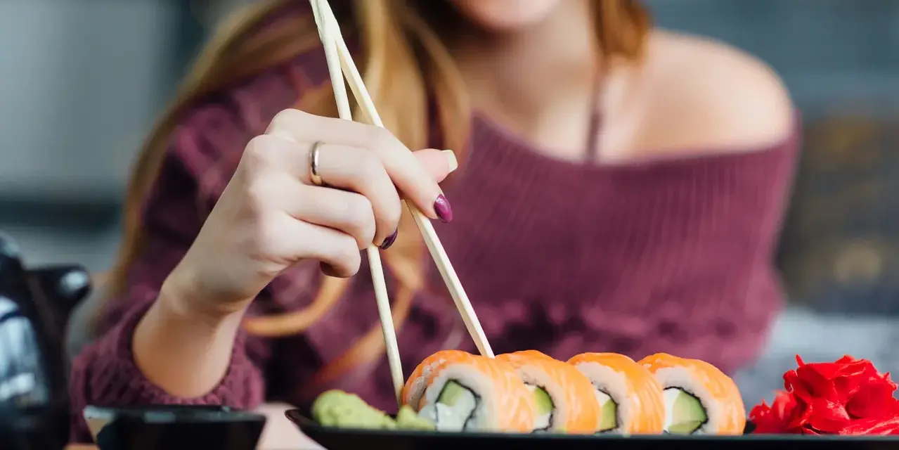 Uśmiechnięta kobieta w ciąży sięga po sushi pałeczkami. Na talerzu łosoś, awokado i imbir.