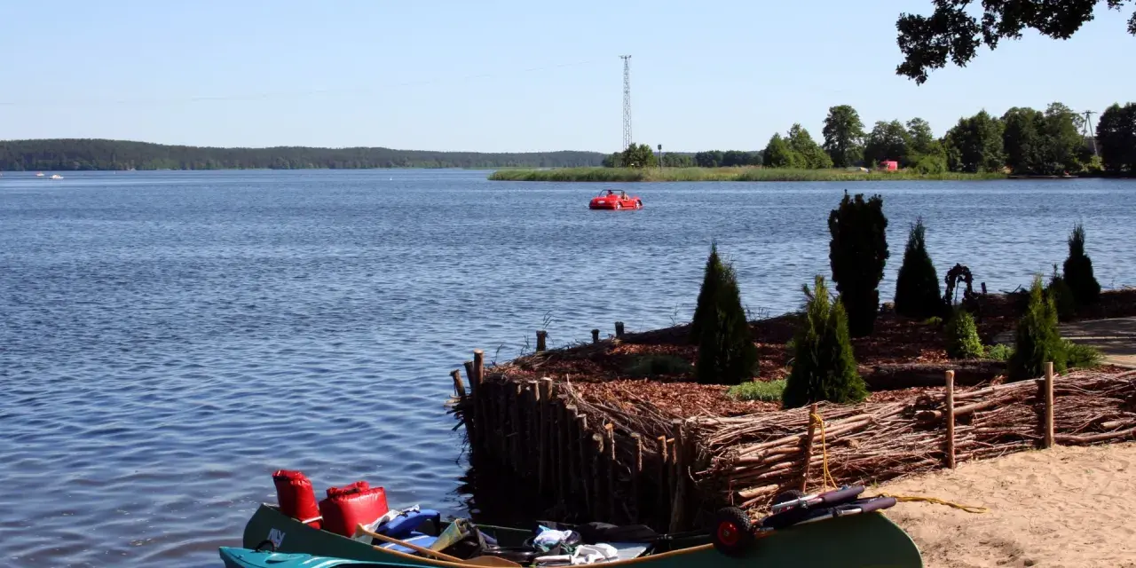 Dwie kajaki na piaszczystej plaży w Swornegaciach na Kaszubach. W oddali czerwona łódka i jezioro.