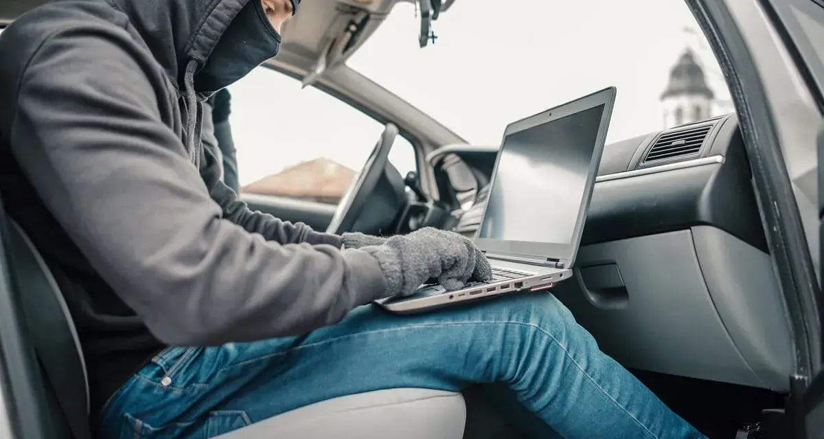 Haker w masce i kapturze próbuje obejść zabezpieczenia keyless go, używając laptopa w samochodzie.
