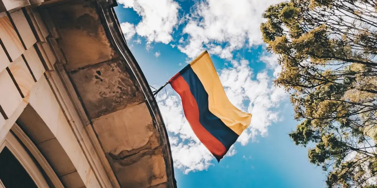 Flaga Kolumbii powiewa na wietrze na tle błękitnego nieba z chmurami, obok zabytkowego budynku i drzew.