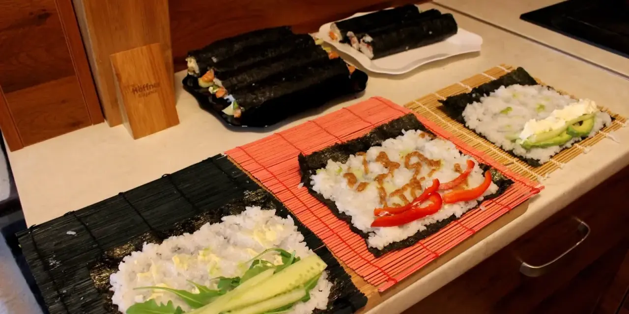Przygotowanie ryżu do sushi. Na matach leżą arkusze nori z ryżem i dodatkami: ogórkiem, awokado, papryką i sosem.
