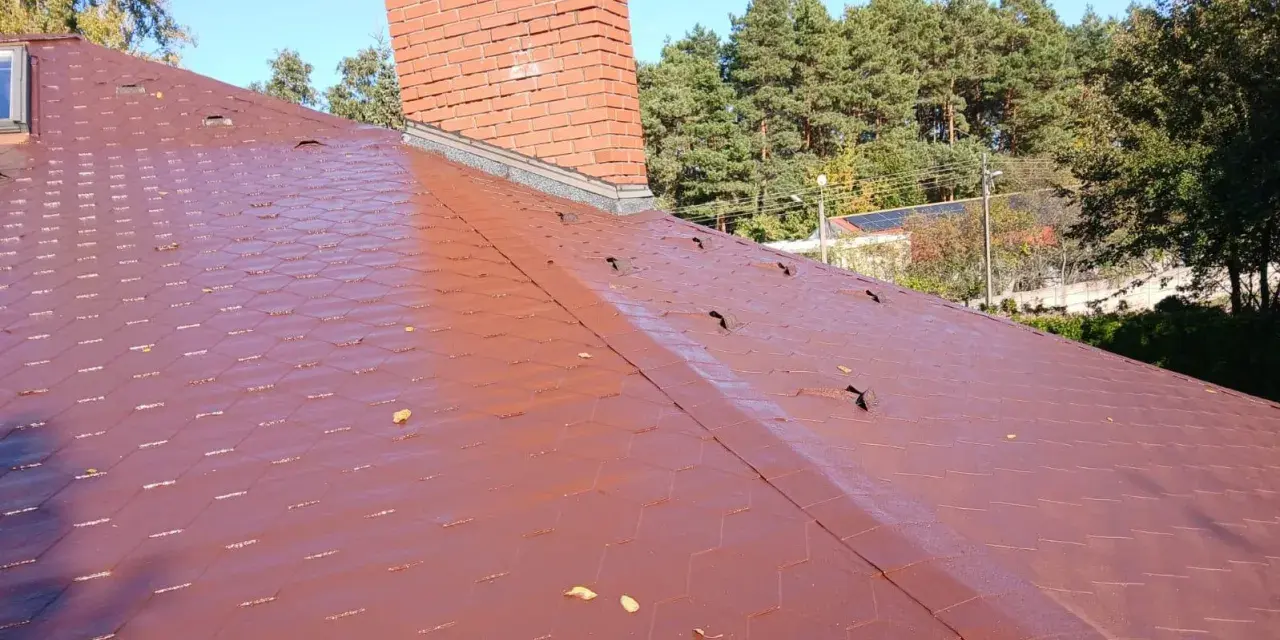Czerwony dach pokryty gontem bitumicznym z widocznym kominem i drzewami w tle.