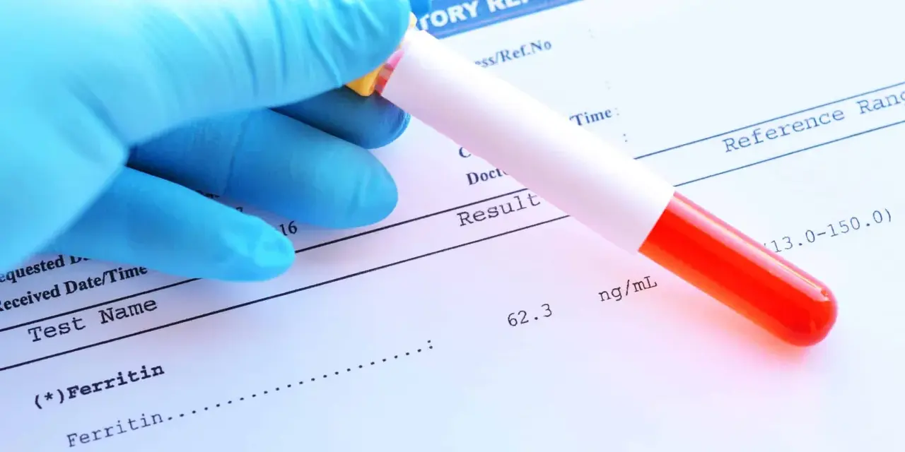 Ręka w niebieskiej rękawiczce trzyma probówkę z krwią nad raportem laboratoryjnym. Wynik testu na **ferratynę** wynosi 62.3 ng/mL.