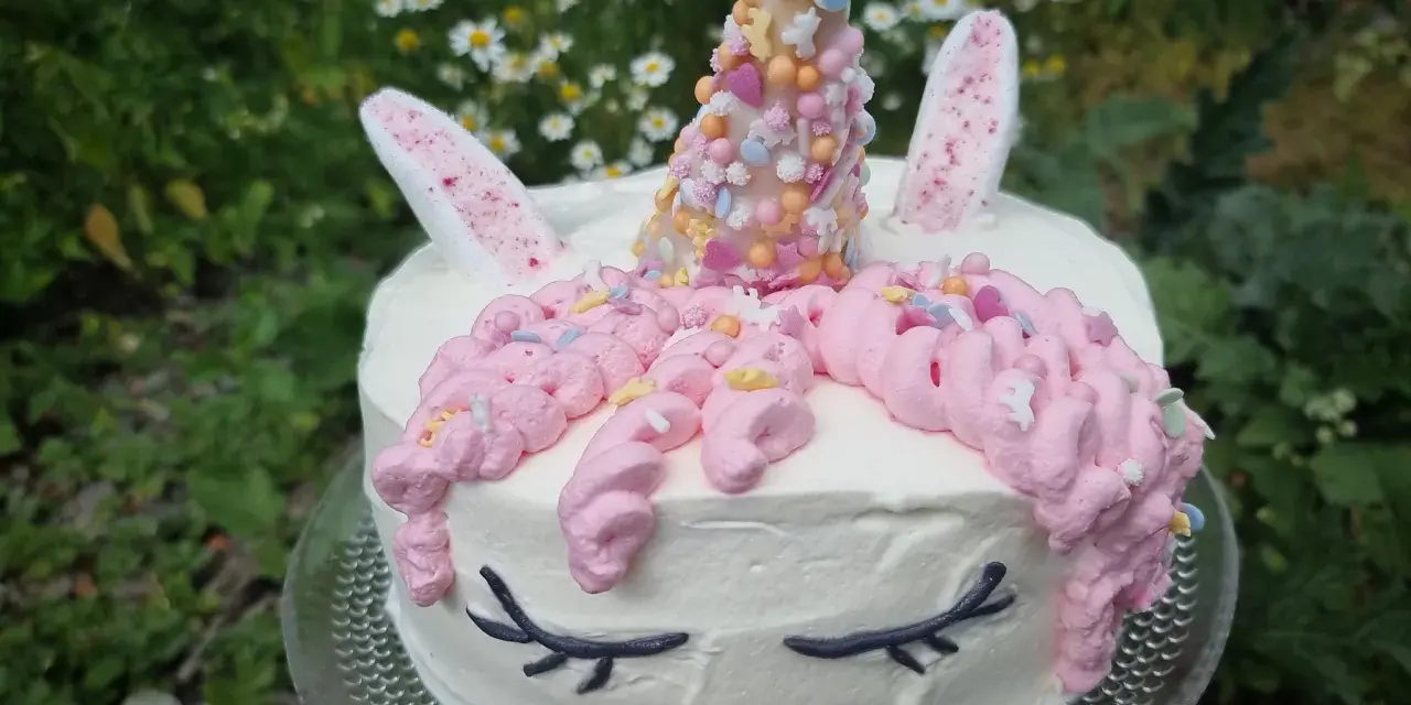 En förtrollande unicorn tårta med rosa glasstrut-liknande horn, dekorerad med strössel och blommor i bakgrunden.