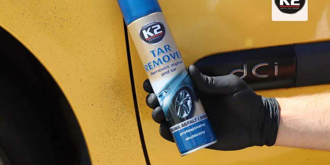 Rękawiczka z czarną gumą trzyma puszkę K2 Tar Remover, która skutecznie usuwa smołę z żółtego lakieru samochodu.