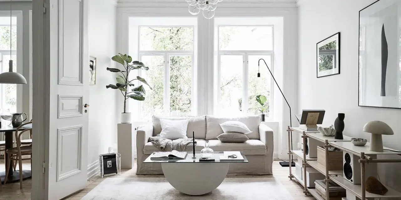 Salón con sofá beige, mesa de centro de cristal y alfombra clara. Decoración minimalista y plantas, creando un ambiente de feng shui.