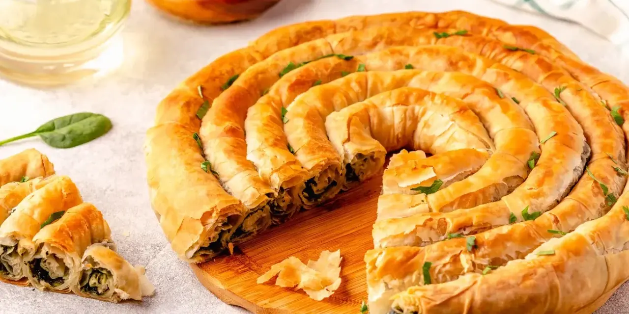 Złocisty borek ze szpinakiem, zwinięty w spiralę, kusi chrupiącym ciastem i aromatycznym nadzieniem. Obok kilka porcji i świeże liście szpinaku.