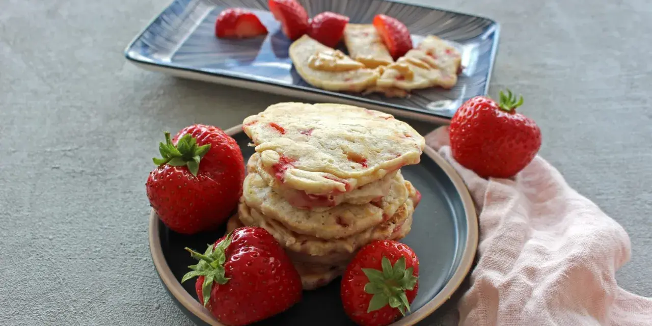 Ein Stapel Erdbeer-Pancakes mit frischen Erdbeeren auf einem Teller. Ein weiterer Teller im Hintergrund zeigt geschnittene Erdbeeren und Pancakes mit Erdnussbutter.