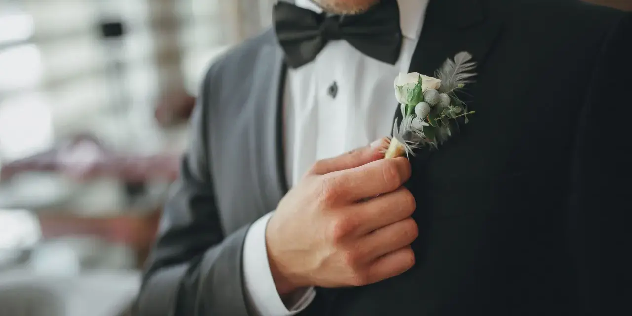 Mężczyzna poprawia butonierkę w eleganckim, czarnym garniturze.