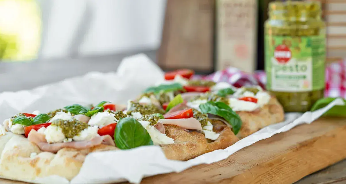 Długi placek z mozzarellą, pomidorkami i bazylią, polany zielonym pesto. Obok słoiczek z pesto.