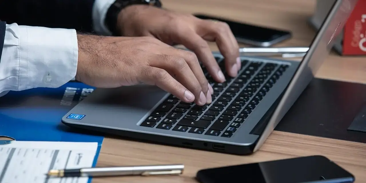 Ręce piszą na laptopie, obok telefon i długopis. Uważaj na oszustów, którzy podszywają się pod polskie firmy.