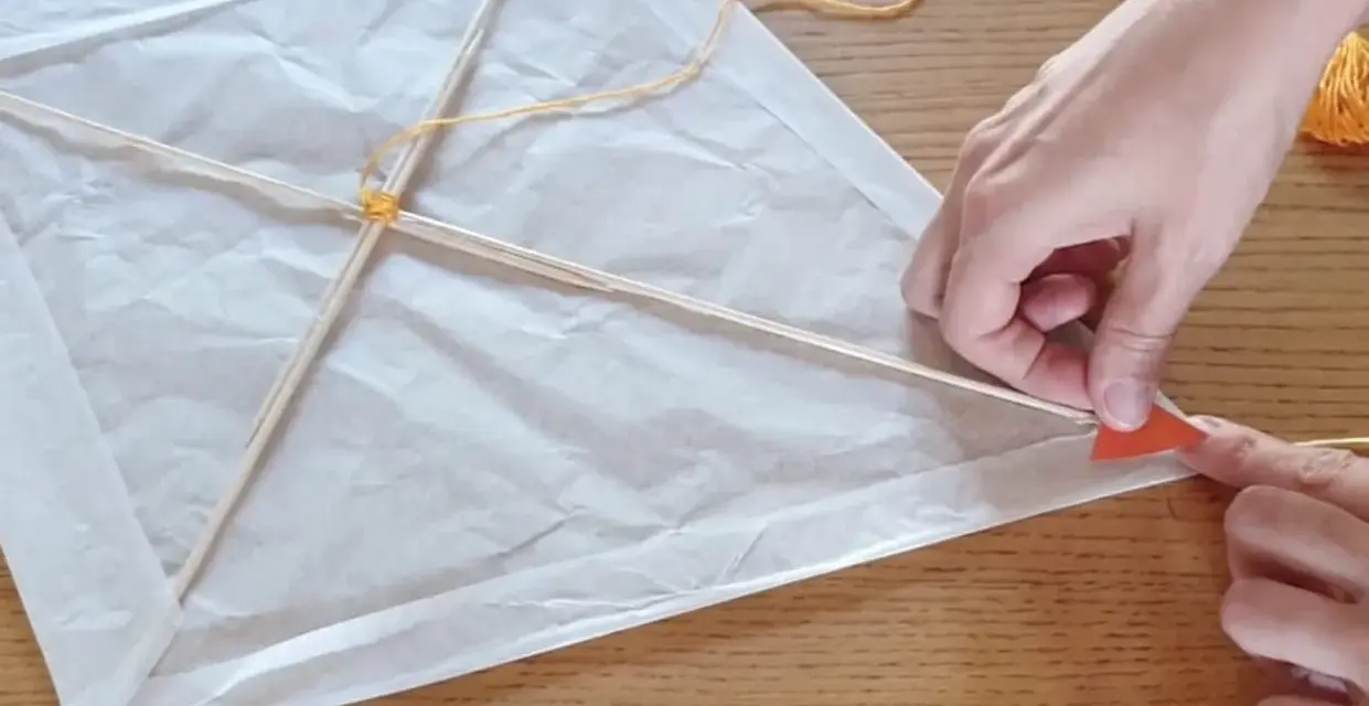 Mains fabriquant un cerf-volant simple en papier blanc et bâtons de bois, avec fil jaune.