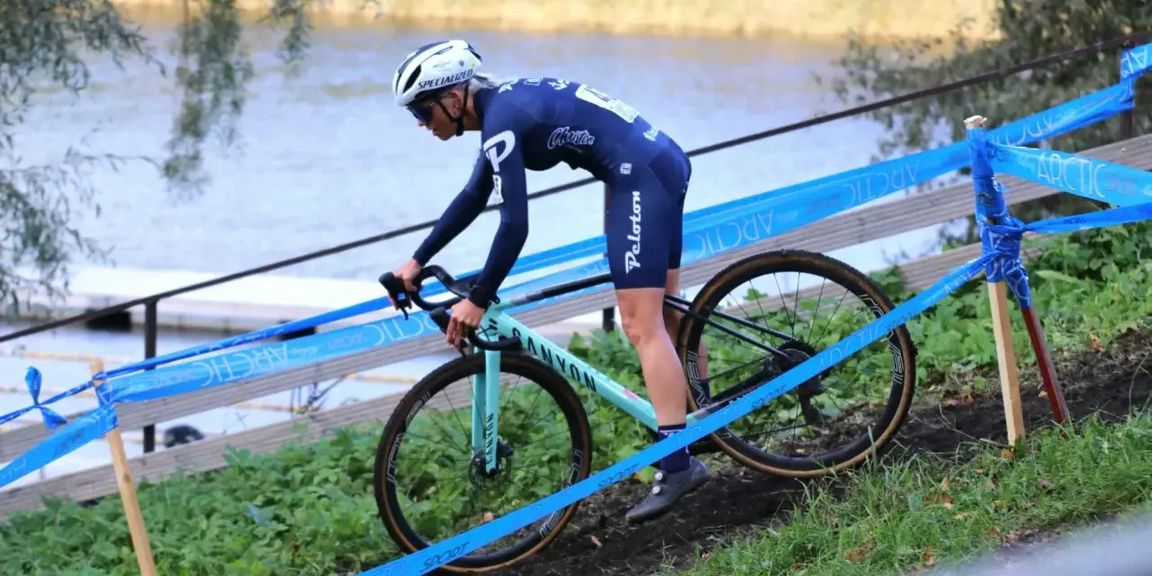Cyclo cross racer in blue kit navigates a muddy course beside a lake, marked by blue tape.