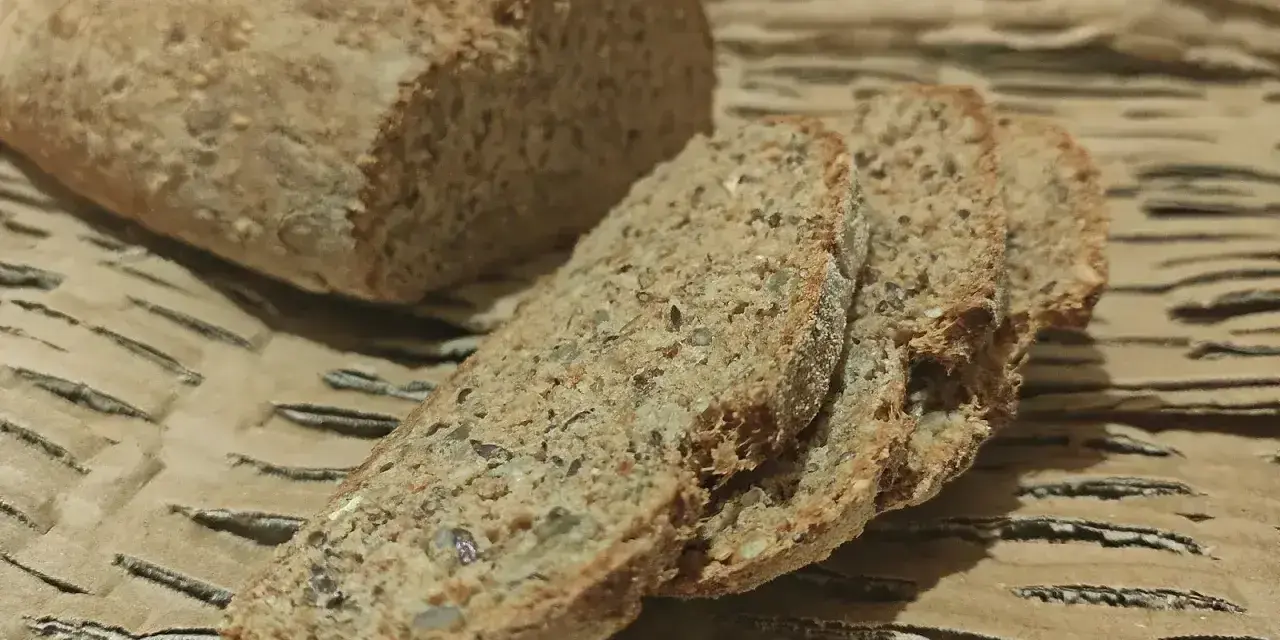 Chleb orkiszowy, pokrojony w kromki, z widocznymi ziarnami, leży na falistej tekturze.