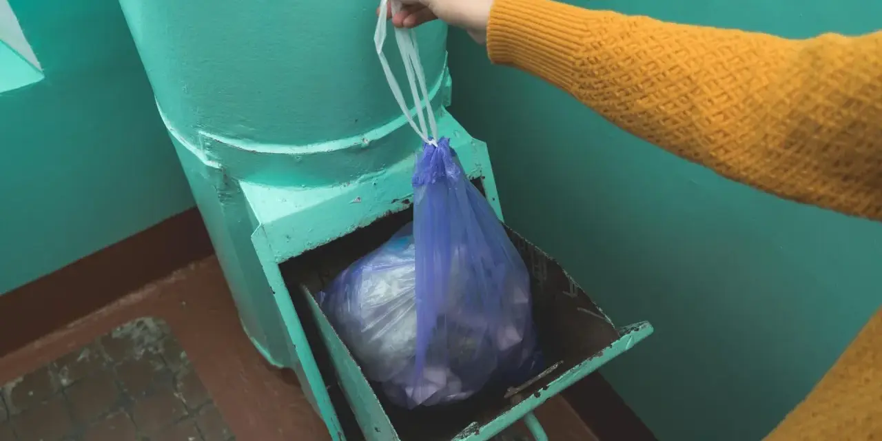 Ręka w żółtym swetrze wkłada worek ze śmieciami do metalowego zsypu w bloku.