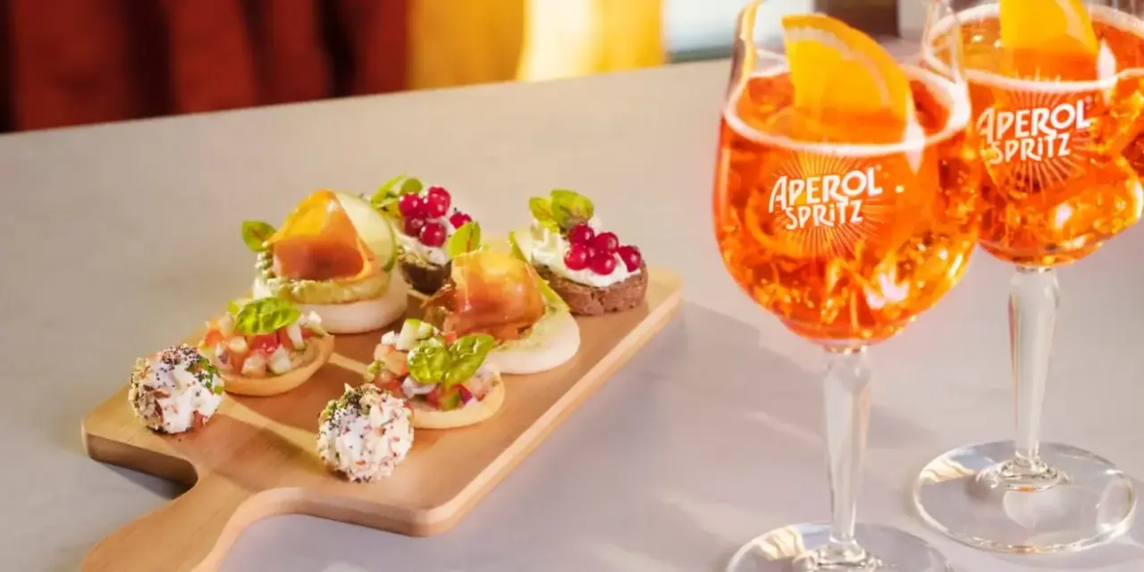 Zwei Aperol Spritz Gläser mit Orangenscheiben und eine Holzplatte mit verschiedenen Häppchen. Perfekt für einen Aperitif!